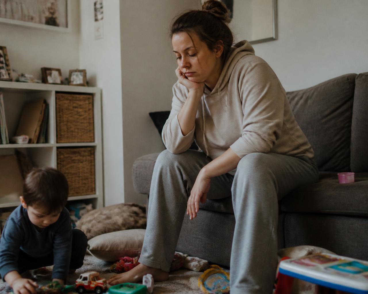 Mãe sentada no sofá no fim do dia, com brinquedos espalhados pelo chão